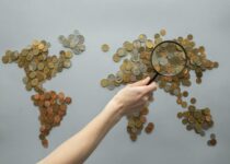 Top view of crop unrecognizable traveler with magnifying glass standing over world map made of various coins on gray background