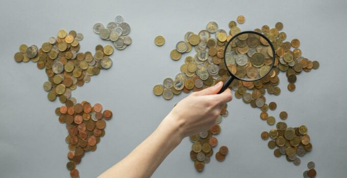 Top view of crop unrecognizable traveler with magnifying glass standing over world map made of various coins on gray background