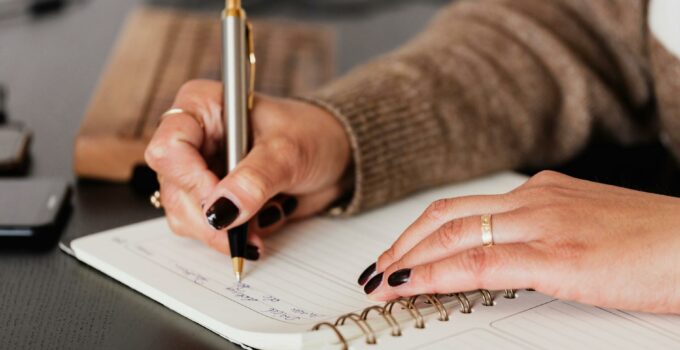 Crop unrecognizable female with stylish manicure sitting at black desk with keyboard and smartphone and taking notes with silver pen in notepad