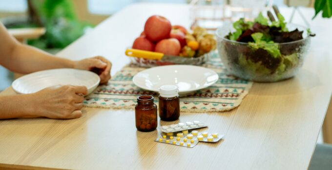 From above of crop anonymous person sitting at table with pile of various medicines and bowls of fresh lettuce salad and ripe fruits in kitchen