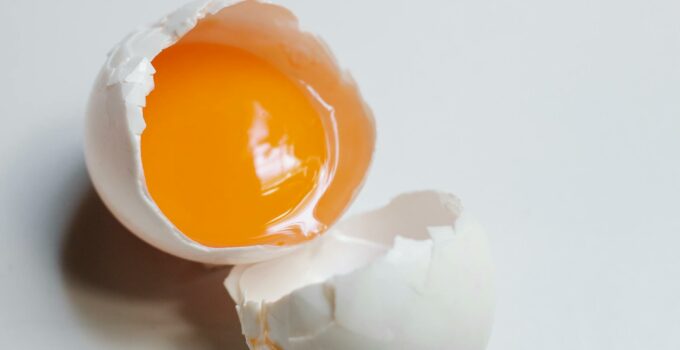 Top view of broken raw egg with yellow yolk and white eggshell on white background in light kitchen during cooking process