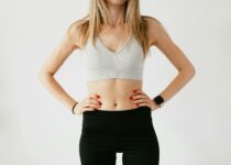 Faceless slim anonymous blond female in sports bra and black leggings in wearable bracelet showing perfect belly on white background while standing with hands on waist