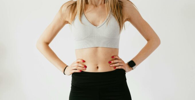 Faceless slim anonymous blond female in sports bra and black leggings in wearable bracelet showing perfect belly on white background while standing with hands on waist