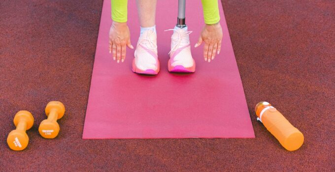 Unrecognizable female athlete with leg prosthesis bending forward and stretching while trying ti touch ground during training on sports ground with dumbbells