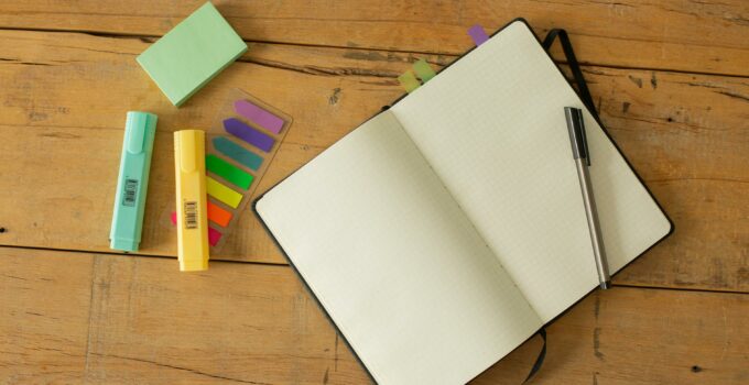 Top view of opened sketchbook with blank pages and pen with coloured markers and bookmarks arranged on wooden desk