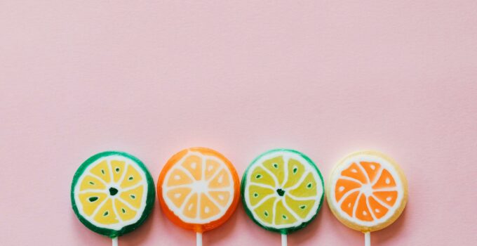 Top view of round multicolored candies with citrus fruit flavor on thin plastic sticks on pale pink surface