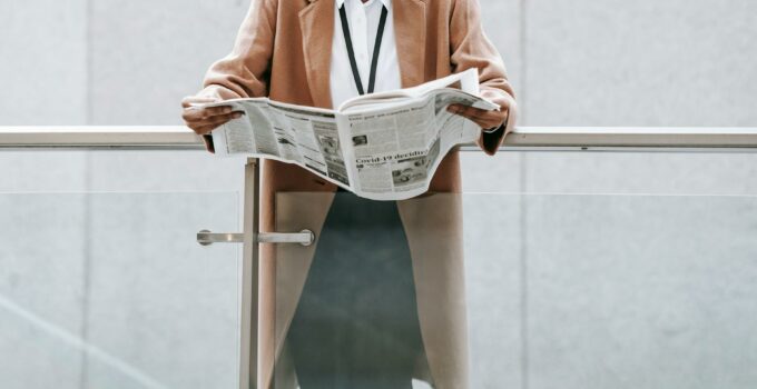 Crop content African American businesswoman in formal clothes reading fresh newspaper and leaning on glass railing of contemporary office building