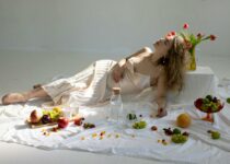 Peaceful young elegant woman lying on floor near fruits scattered on white cloth