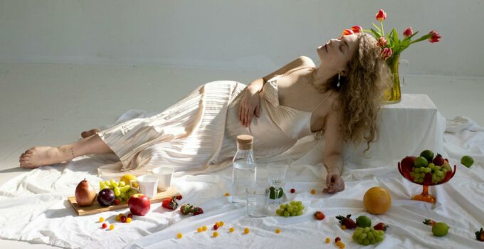 Peaceful young elegant woman lying on floor near fruits scattered on white cloth