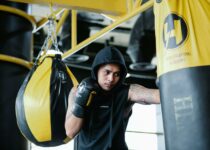 Focused young Latin American tattooed male fighter in hooded activewear and boxing gloves hitting punching bag during training in sports club