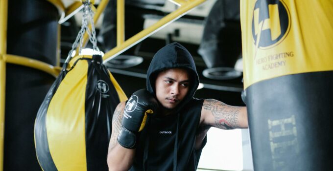 Focused young Latin American tattooed male fighter in hooded activewear and boxing gloves hitting punching bag during training in sports club