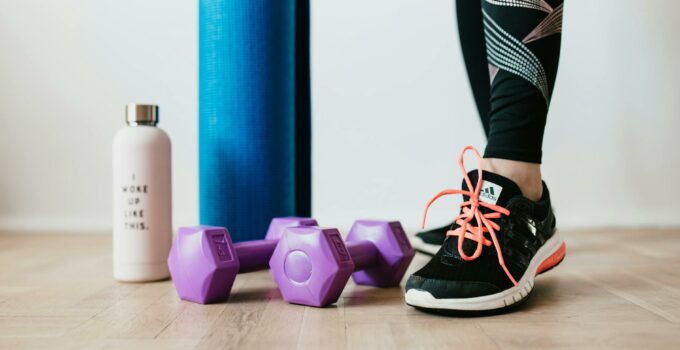 Crop sportswoman wearing sneakers and black leggings standing with sport mat on wooden floor near dumbbells and water bottle before exercising indoors