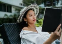 Focused young ethnic lady in white shirt and straw hat lying on sunbed in hotel yard and reading information in notebook