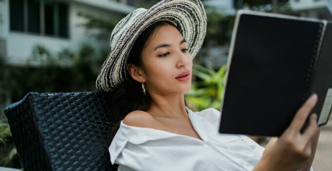 Focused young ethnic lady in white shirt and straw hat lying on sunbed in hotel yard and reading information in notebook