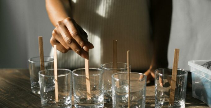 Unrecognizable African American female securing wooden wicks in glass molds while making aroma candles at wooden table in light studio