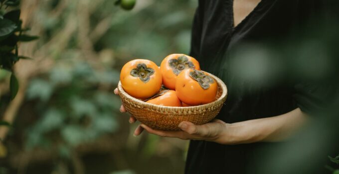 Crop faceless female in black dress with wicker bowl full of ripe fresh fruits of date plums