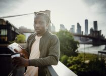 Wistful black student standing on balcony with coffee and notebook