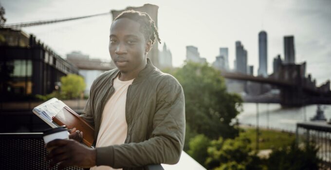 Wistful black student standing on balcony with coffee and notebook