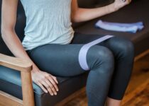 Faceless slim strong female in sportswear sitting on couch while exercising with elastic resistance band on legs during intense workout
