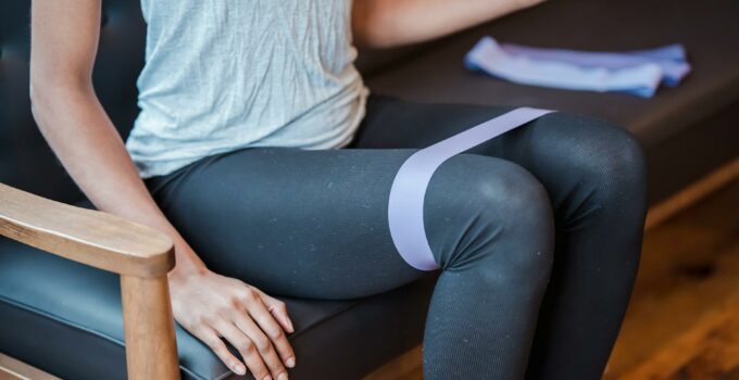 Faceless slim strong female in sportswear sitting on couch while exercising with elastic resistance band on legs during intense workout